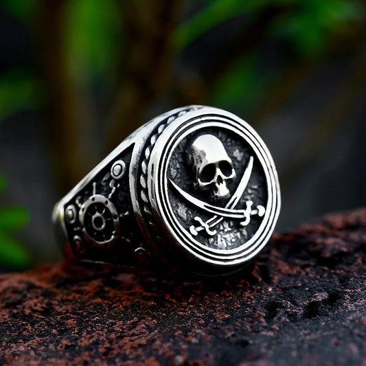 Front view of The Captain's Oath stainless steel ring, featuring a skull with crossed swords engraved on its face, set on a textured surface.