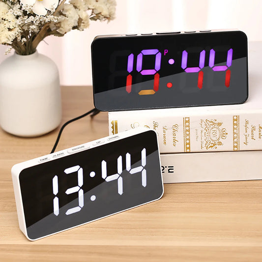 Two digital alarm clocks, black and white, on a desk with books, displaying different LED time colors.