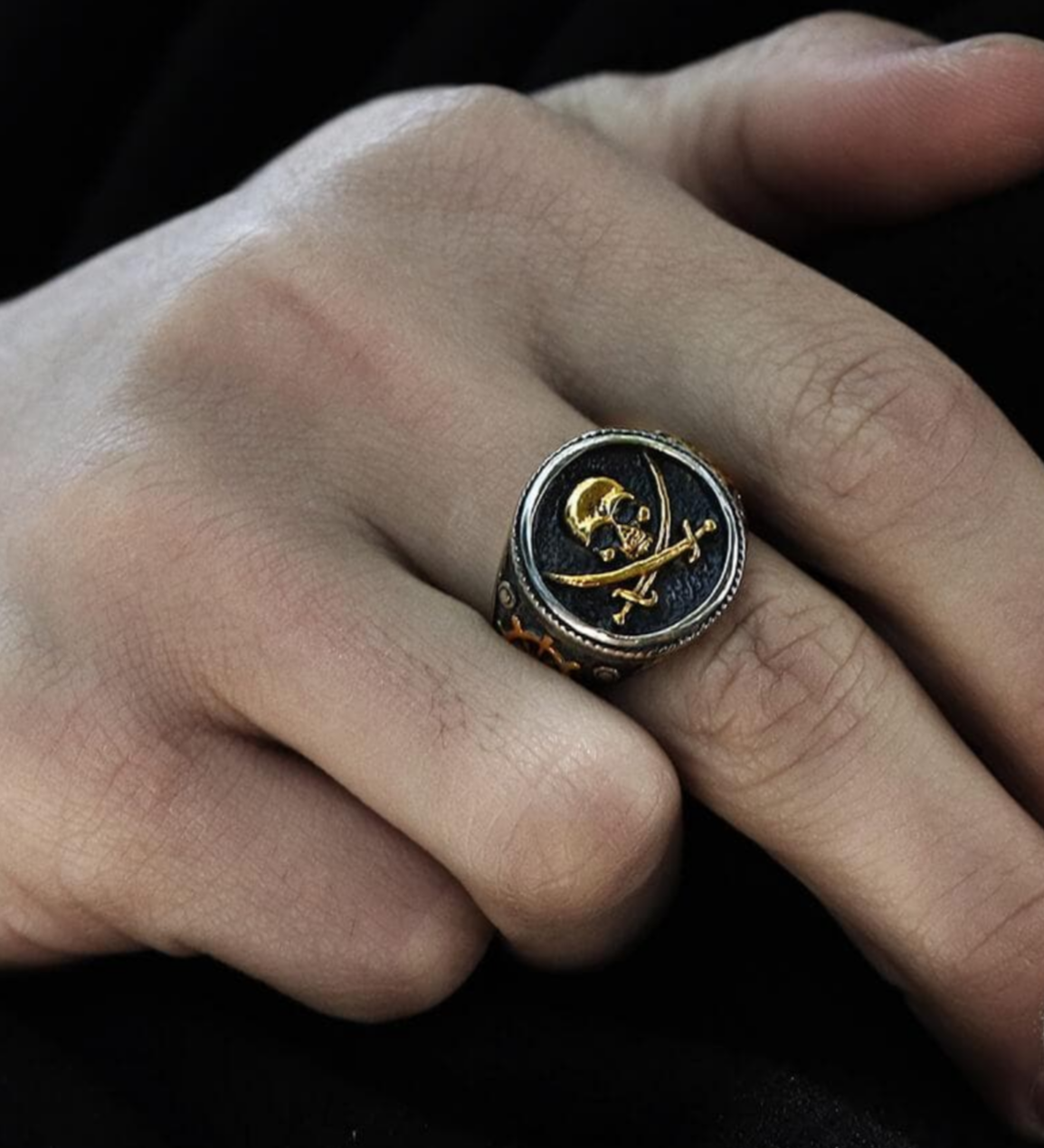 Close-up of a pirate ring with a gold skull and crossbones on a person's finger, showing intricate side detailing.