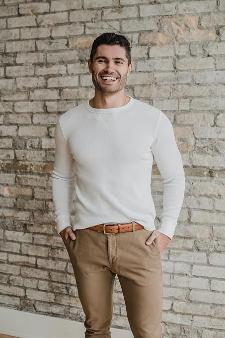 A smiling man wearing a white long-sleeve crewneck knit sweater and tan pants, standing against a light brick wall.